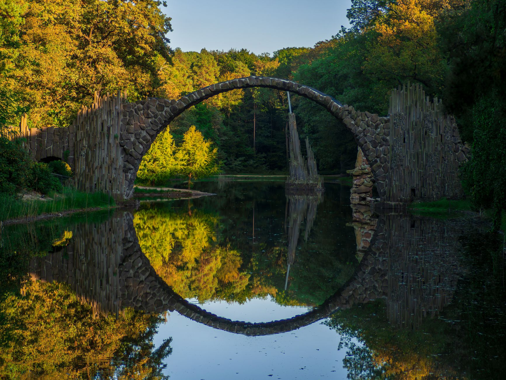 Photo by Atlas on Pexls.com, A curved bridge mirrored in the water below, fall leaves.